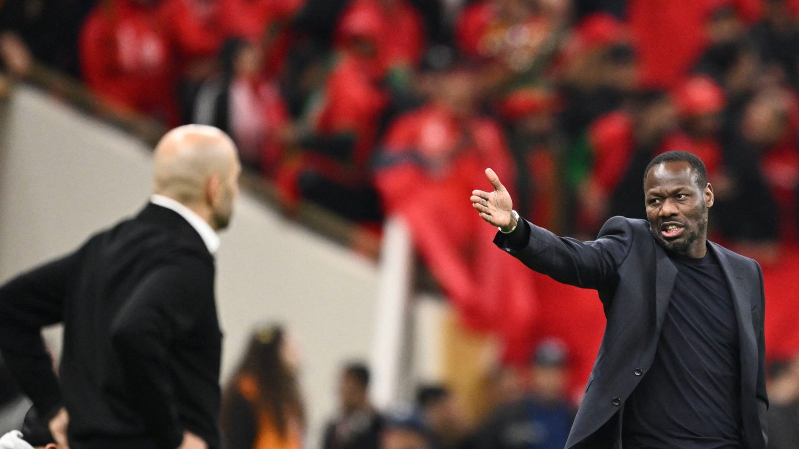 Twee voetbalcoaches in gesprek aan de zijlijn, waarbij één gepassioneerd gebaren maakt. Marokkaanse fans in rode kleding vullen de achtergrond en creëren een elektriserende sfeer.