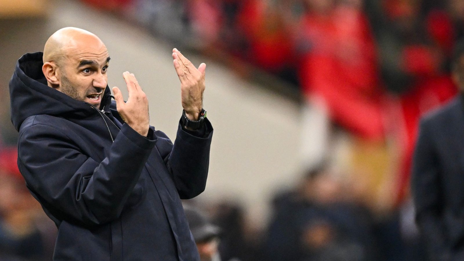 Walid Regragui gesticule avec animation en bord de terrain, portant un manteau marine, lors d'un moment tendu du match, tandis que des supporters en rouge acclament en arrière-plan.