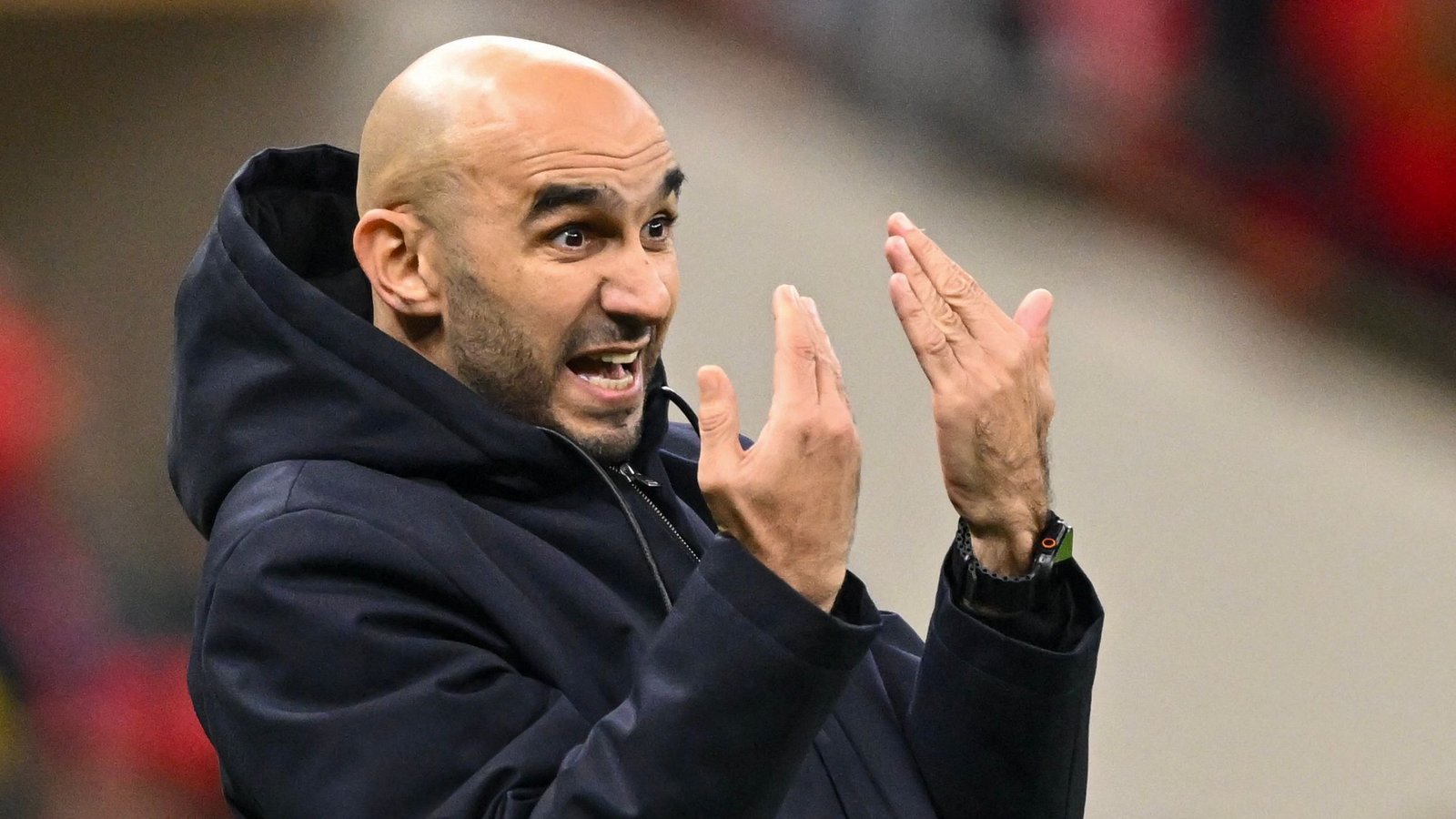 Walid Regragui gesticule avec passion en bord de terrain, vêtu d'un sweat à capuche sombre, lors d'un moment intense d'un match de football marocain.