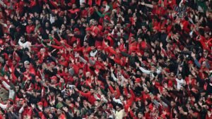 Les supporters marocains, vêtus de rouge et vert, célèbrent avec passion en agitant les bras dans une atmosphère de stade vibrante lors d'un match de football.
