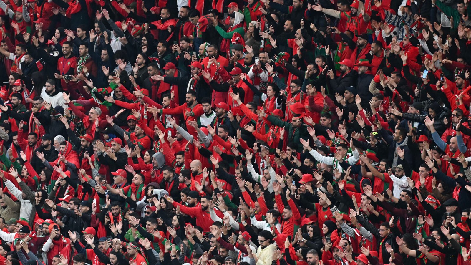 Les supporters marocains, vêtus de rouge et vert, célèbrent avec passion en agitant les bras dans une atmosphère de stade vibrante lors d'un match de football.