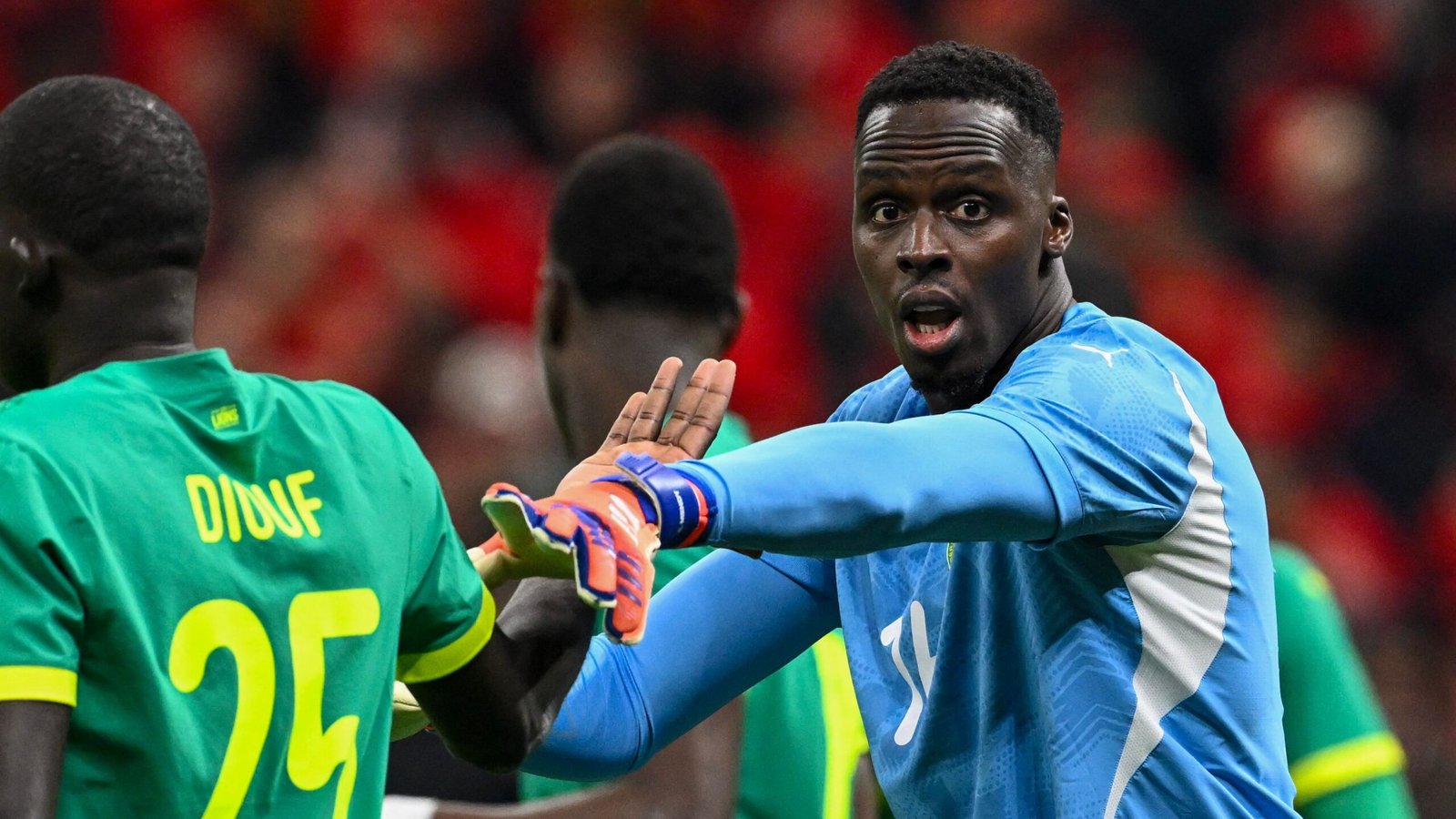 Senegal goalkeeper gestures passionately during a match, wearing blue gloves and a kit, surrounded by teammates in green.