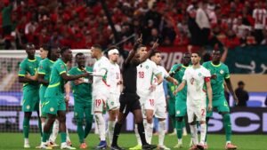 Les joueurs marocains et sénégalais se rassemblent autour de l'arbitre pendant un moment tendu, arborant les couleurs d'équipe vert et blanc, avec des supporters d'un rouge vif.