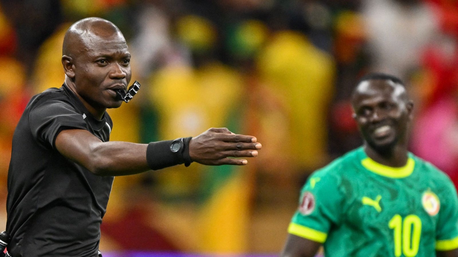 L'arbitre fait un geste pendant un match, avec un joueur sénégalais souriant en tenue verte, devant un fond de foule animée.