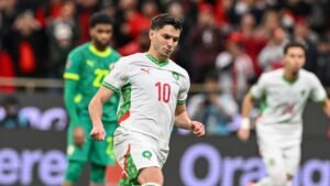 Youssef En-Nesyri in Morocco's white and green kit, preparing for a play during a match against Senegal, with excited fans in the background.