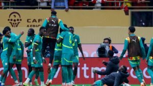 Senegal players celebrate a goal during a CAF Africa Cup of Nations match, showcasing their teal and yellow kits against a lively crowd.