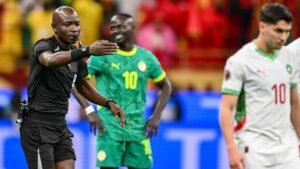 L'arbitre fait des gestes pendant un match, encadré par le joueur sénégalais en vert et le joueur marocain numéro 10, dans une atmosphère de foule vibrante.