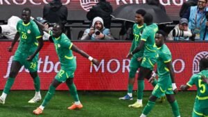 Senegal players in vibrant green kits celebrate a goal, showing excitement against a backdrop of fans and a match photographer.