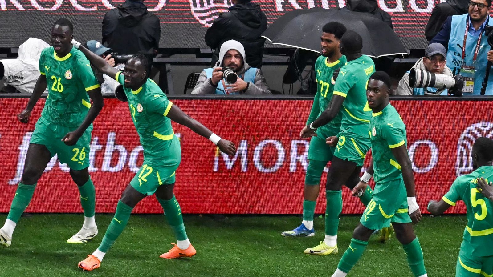 Senegal players in vibrant green kits celebrate a goal, showing excitement against a backdrop of fans and a match photographer.