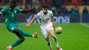 Moroccan player wearing jersey number 17 challenges a Senegalese defender in heavy rain, with a football in play on a vibrant green pitch.