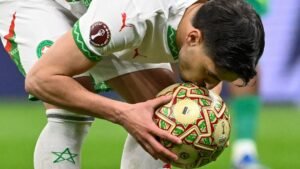 Un joueur marocain embrasse le ballon, portant un maillot blanc et vert, le logo de la Coupe d'Afrique des Nations étant visible sur le ballon.