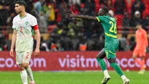 لاعب سنغالي يحتفل بفرح بالزي الأخضر، بينما يبدو أشرف حكيمي المغربي، باللون الأبيض، محبطًا وسط ظروف مباراة ممطرة.