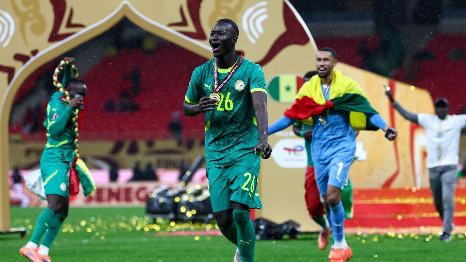 Senegalese speler viert met een gouden medaille, gekleed in een groen tenue versierd met kleurrijke accessoires, terwijl teamgenoten op de achtergrond met vlaggen zwaaien.