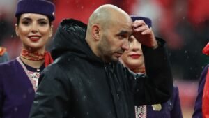 Walid Regragui, l'air pensif dans une veste noire, encadré par des femmes souriantes et joyeuses vêtues de tenues violettes et rouges, sur fond de pluie.