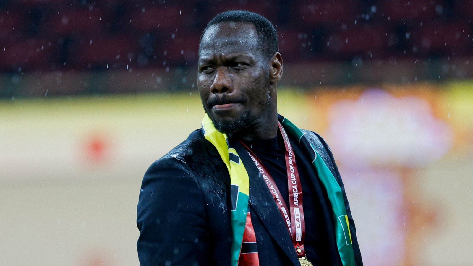 Feest in de regen, een trotse coach in een zwart pak en medaille, gehuld in een groen-gele vlag, toont emotie na een wedstrijd.