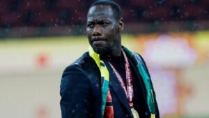 مدرب كرة قدم يحتفل تحت المطر مرتدياً بدلة سوداء وألواناً وطنية، حاملاً ميدالية كأس الأمم الأفريقية.