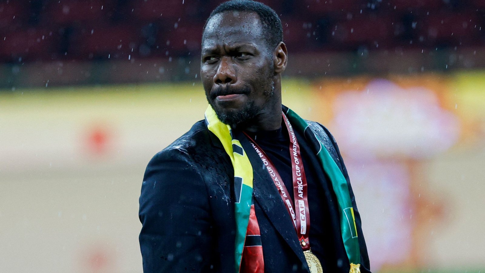 مدرب كرة قدم يحتفل تحت المطر مرتدياً بدلة سوداء وألواناً وطنية، حاملاً ميدالية كأس الأمم الأفريقية.