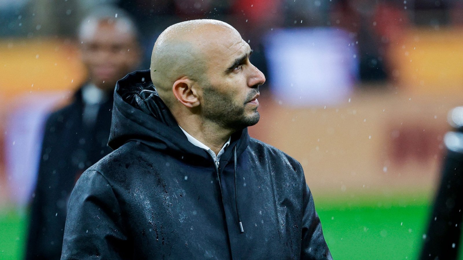 Walid Regragui, de bondscoach van Marokko, staart peinzend in de regen, gekleed in een donker jasje, wat een aangrijpend moment vastlegt.