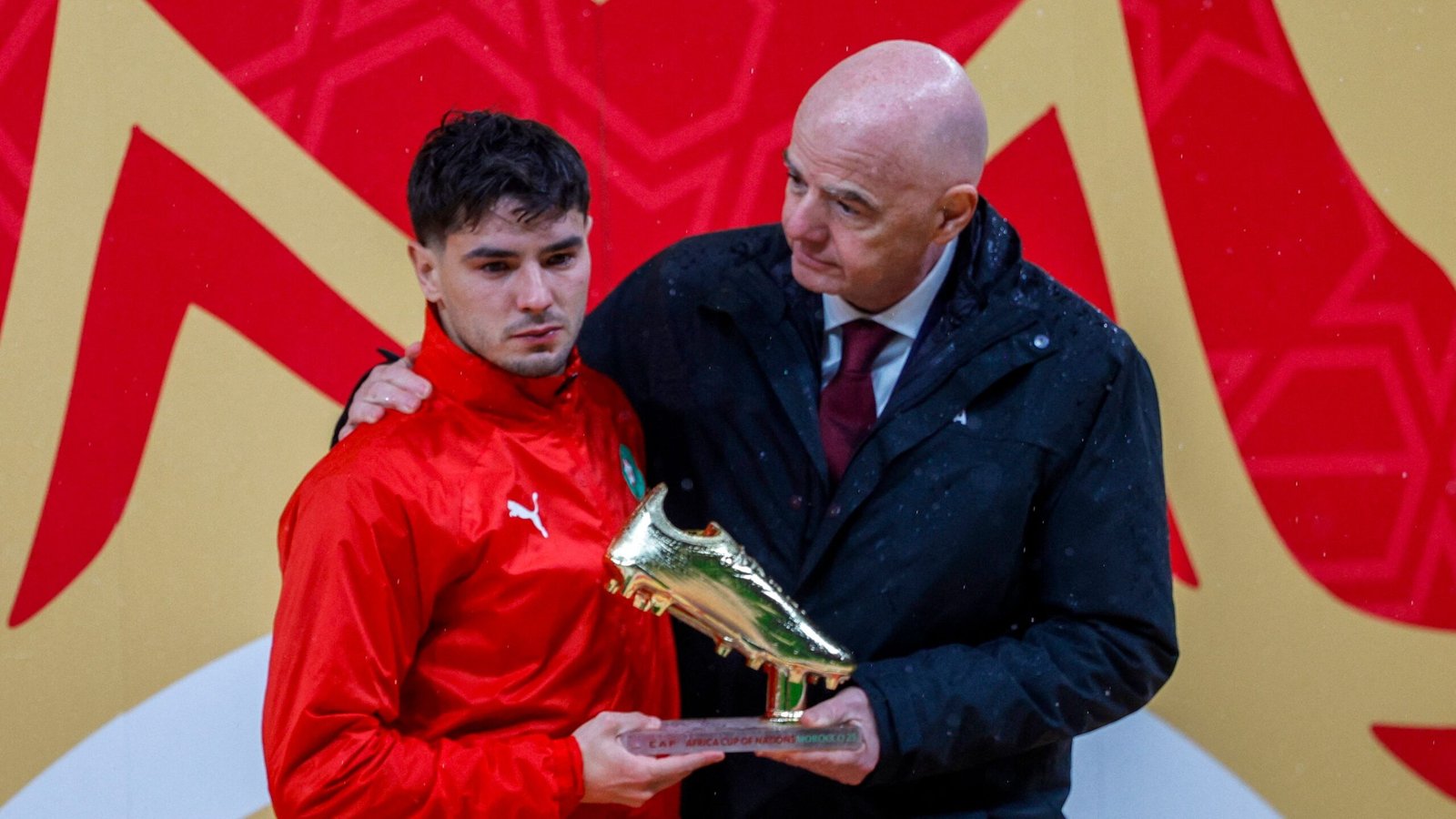 Moroccan player receiving a golden boot trophy, standing under a rain-kissed backdrop, with a dignitary offering congratulations.