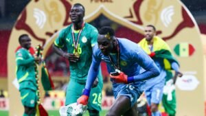 Les joueurs sénégalais célèbrent leur victoire à la Coupe d'Afrique des Nations, le gardien de but en bleu tenant un trophée avec ses coéquipiers en maillots verts derrière lui.