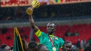 Sadio Mané lifts the African Cup of Nations trophy, celebrating with teammates in a rain-soaked stadium.