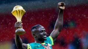 Sadio Mané celebrates holding the Africa Cup trophy, wearing a turquoise national team kit, with a jubilant expression in the rain.