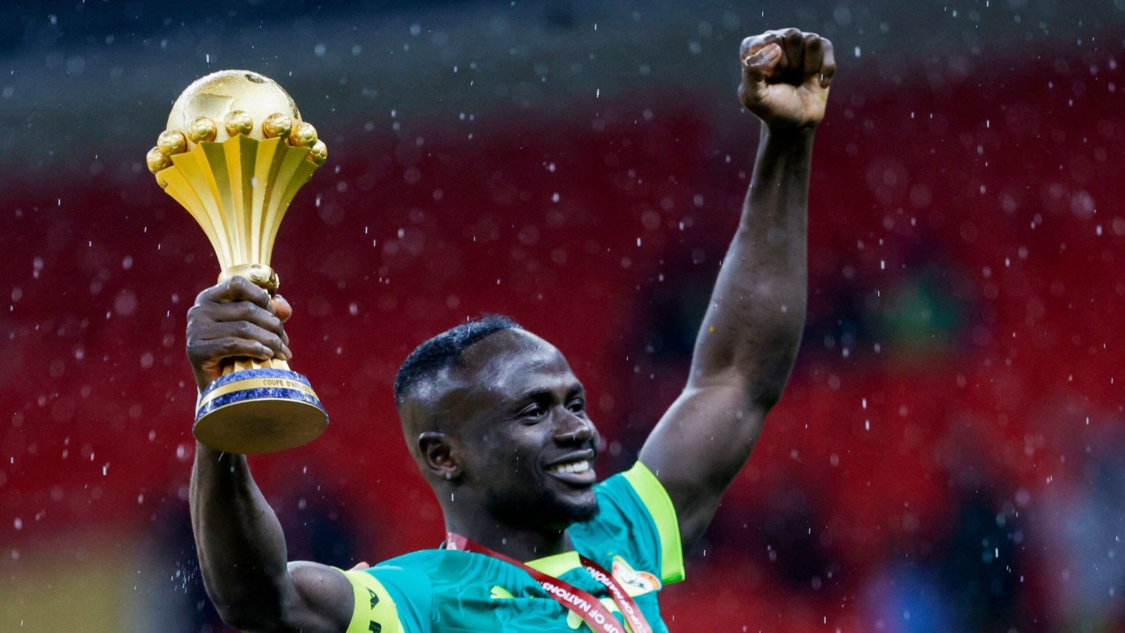 Sadio Mané celebrates holding the Africa Cup trophy, wearing a turquoise national team kit, with a jubilant expression in the rain.