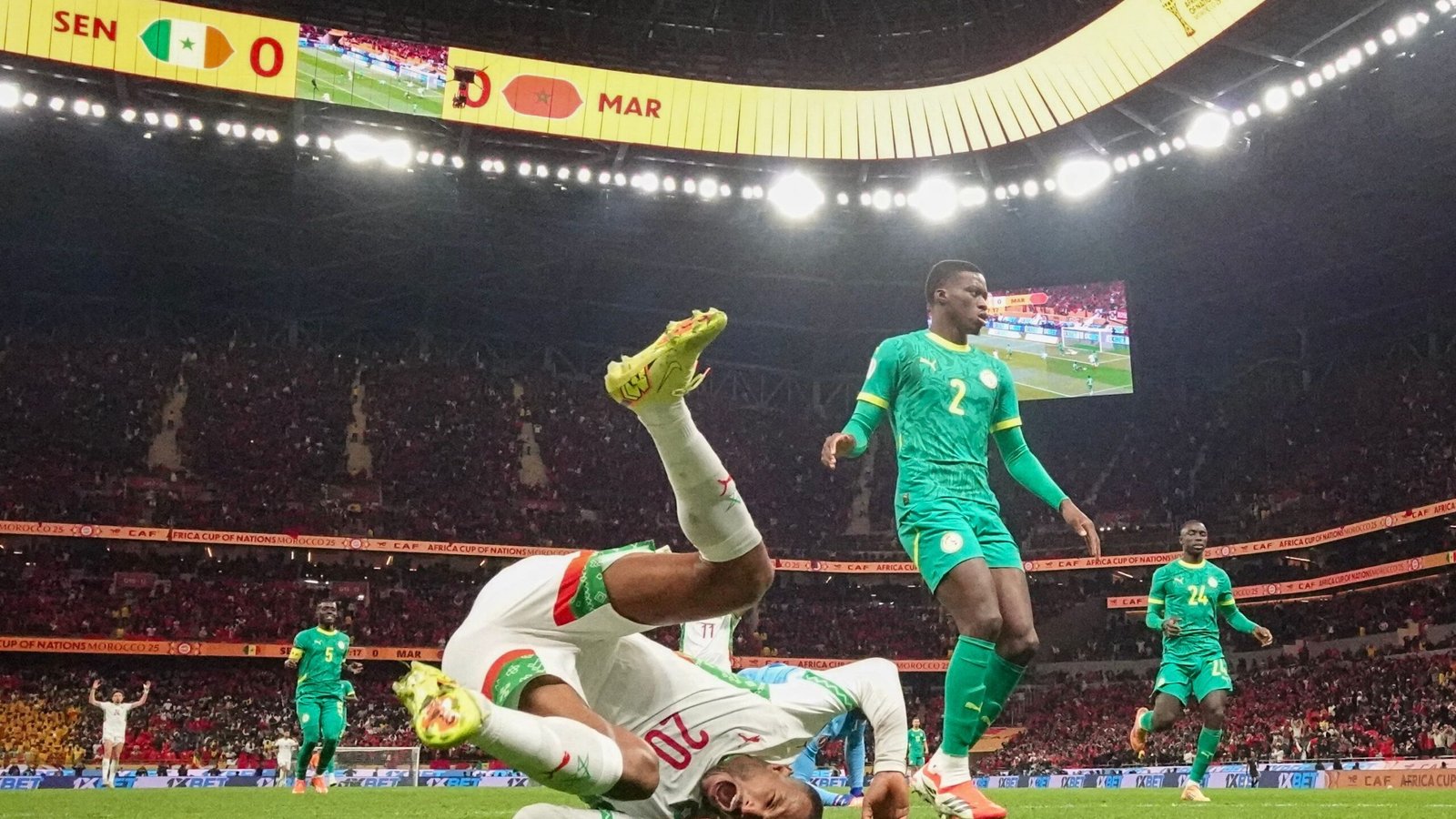 Moroccan player falls to the ground as Senegalese defenders stand nearby, with a scoreboard showing Senegal 0 - 0 Morocco in a vibrant stadium.