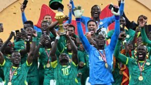 Senegal national team celebrates holding the Africa Cup trophy aloft, surrounded by joyous players in green jerseys, gold medals, and confetti.