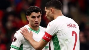 أشرف حكيمي يتشارك لحظة مع زميله في الفريق، وكلاهما يرتديان الطقم الأبيض للمغرب بتطريزات خضراء خلال موقف مباراة متوتر.