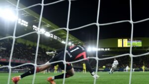 Speler van Real Madrid neemt een strafschop terwijl de doelman van Villarreal duikt, met stadionverlichting en een scorebord op de achtergrond.