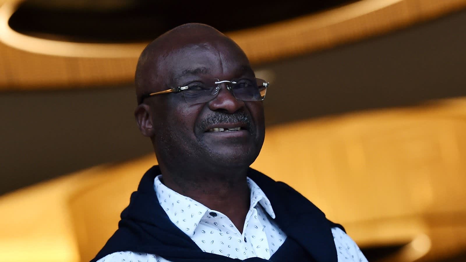 Un homme souriant portant des lunettes et une chemise blanche, avec un pull bleu drapé sur les épaules, se tient devant un fond à l'éclairage doux.