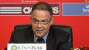 M. Fouzi Lekjaa speaks at a press conference, seated behind a table with a nameplate, against a vibrant red background featuring logos.