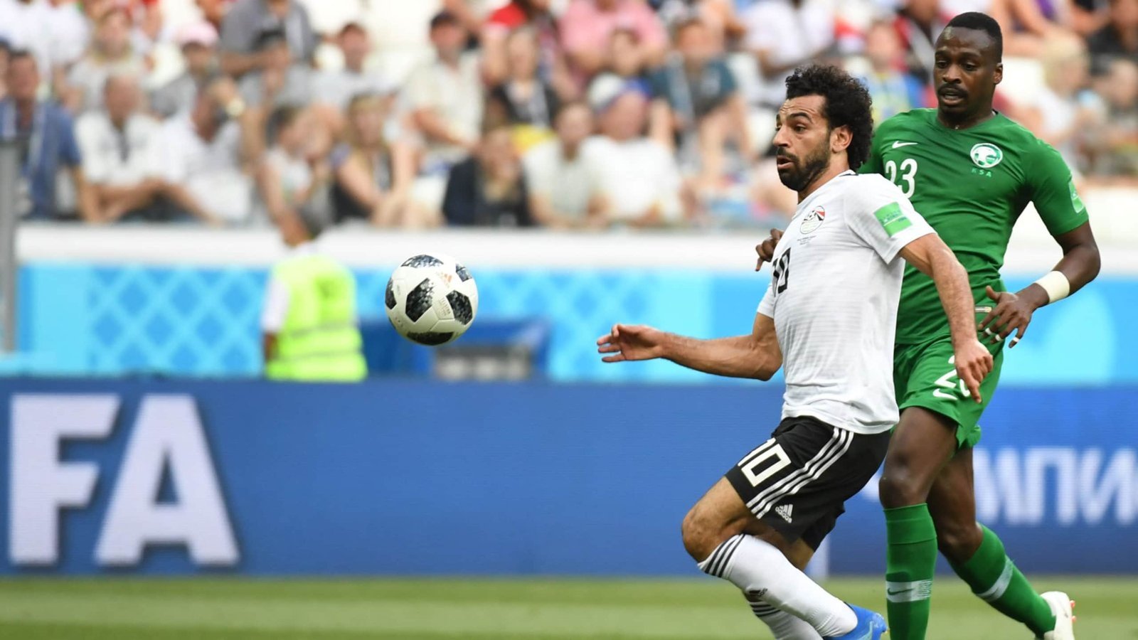 Mohamed Salah in Egypt's white kit faces pressure from a Saudi Arabian defender in green, with a football airborne in a World Cup match.