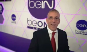 Hafid Derradji smiles confidently in front of a stylish beIN Sports backdrop, wearing a black suit and red tie.