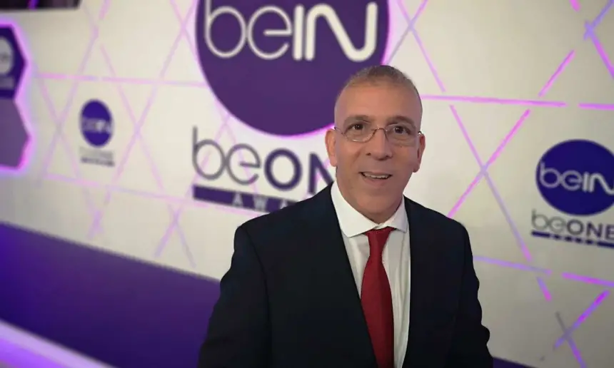 Hafid Derradji smiles confidently in front of a stylish beIN Sports backdrop, wearing a black suit and red tie.
