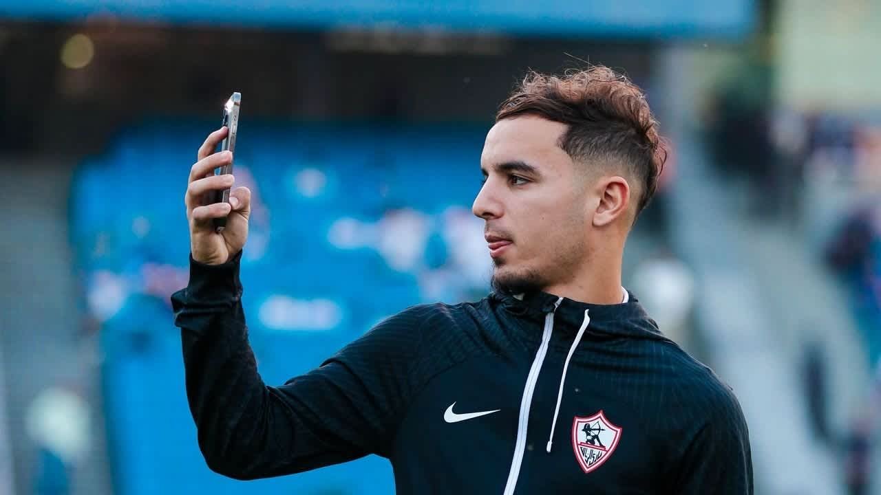 لاعب الزمالك يلتقط صورة ذاتية مرتدياً سترة التدريب السوداء بشعار النادي، مع إبراز خلفية الملعب وأجواء المباراة.
