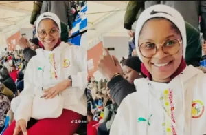Un fan en train de crier dans une veste blanche avec le logo de l'équipe nationale du Sénégal sourit avec enthousiasme dans un stade vibrant rempli de supporters.