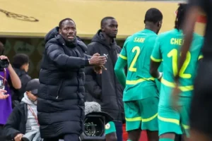 Le sélectionneur du Sénégal fait des gestes depuis la touche tandis que les joueurs Sarr et Cheikha, vêtus de leurs tenues vert vif, s'apprêtent à réintégrer le match.