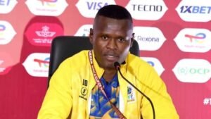 Press conference with a football coach in a yellow jersey, speaking into a microphone, against a vibrant red backdrop.