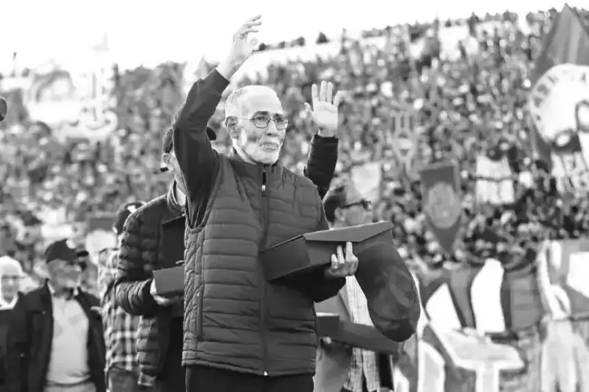 A senior man in a black jacket waves to a cheering crowd while holding a box, amidst a backdrop of colourful banners and fans.