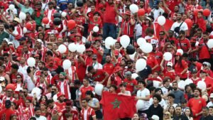 Marokkaanse fans juichen enthousiast in levendige rode en witte kleding, zwaaiend met ballonnen en een vlag, terwijl ze hun team aanmoedigen tijdens een wedstrijd.