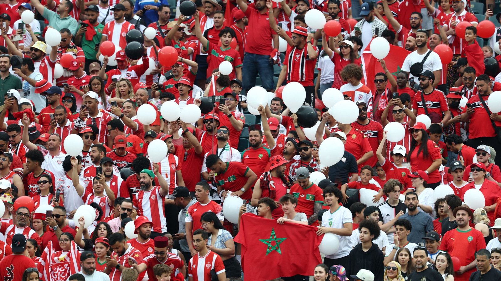 Marokkaanse fans juichen enthousiast in levendige rode en witte kleding, zwaaiend met ballonnen en een vlag, terwijl ze hun team aanmoedigen tijdens een wedstrijd.