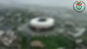 Aerial view of a large, modern football stadium surrounded by green spaces and city infrastructure, set against a cloudy sky.
