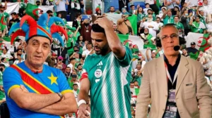 Riyad Mahrez, en tenue verte de l'Algérie, semble abattu, encadré par un supporter portant un chapeau coloré et un commentateur avec un casque, des drapeaux étant visibles dans la foule.