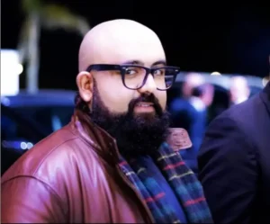 Homme barbu portant des lunettes, vêtu d'une veste en cuir marron et d'une écharpe, capturé dans un environnement à faible luminosité, avec des silhouettes floues en arrière-plan.