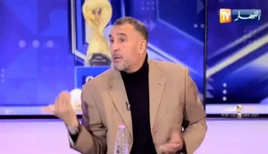 محلل كرة القدم يناقش التكتيكات في برنامج رياضي، يرتدي بلازراً بُنياً فاتحاً أمام خلفية زرقاء، مع ظهور كأس العرب في المشهد.