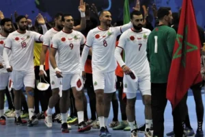Moroccan handball team players in white jerseys celebrate with flags, showcasing team spirit after a match, against a blue backdrop.
