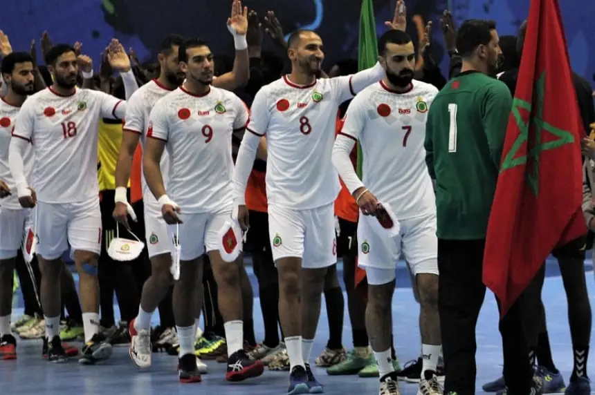 Moroccan handball team players in white jerseys celebrate with flags, showcasing team spirit after a match, against a blue backdrop.