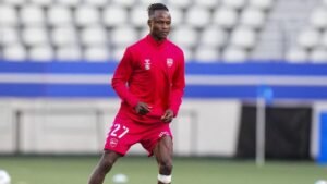 التعاقد الأفريقي الجديد لنادي الرجاء البيضاوي يرتدي زي تدريب أحمر، ويظهر رشاقة في الملعب مع خلفية ملعب حديث.